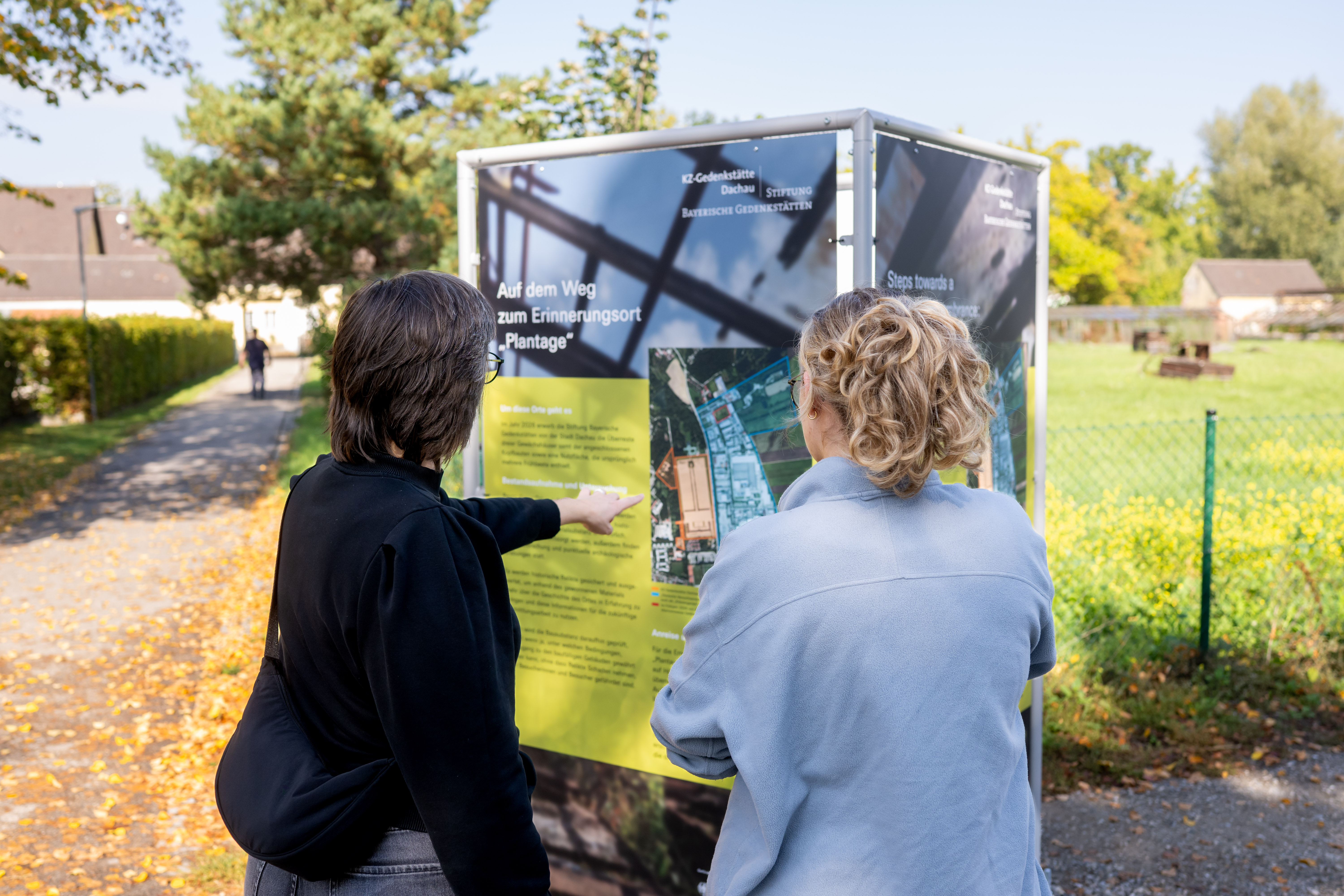 Fotografie zweier Personen vor einer Informationstafel im Außenbereich, die über einen Weg zum Erinnerungsort "Plantage" informiert. Tafel zeigt Fotos, Lagekarte, weiße Beschriftungen und gelben Informationstext; Umgebung mit Gehweg, Bäumen und Wohnhäusern.