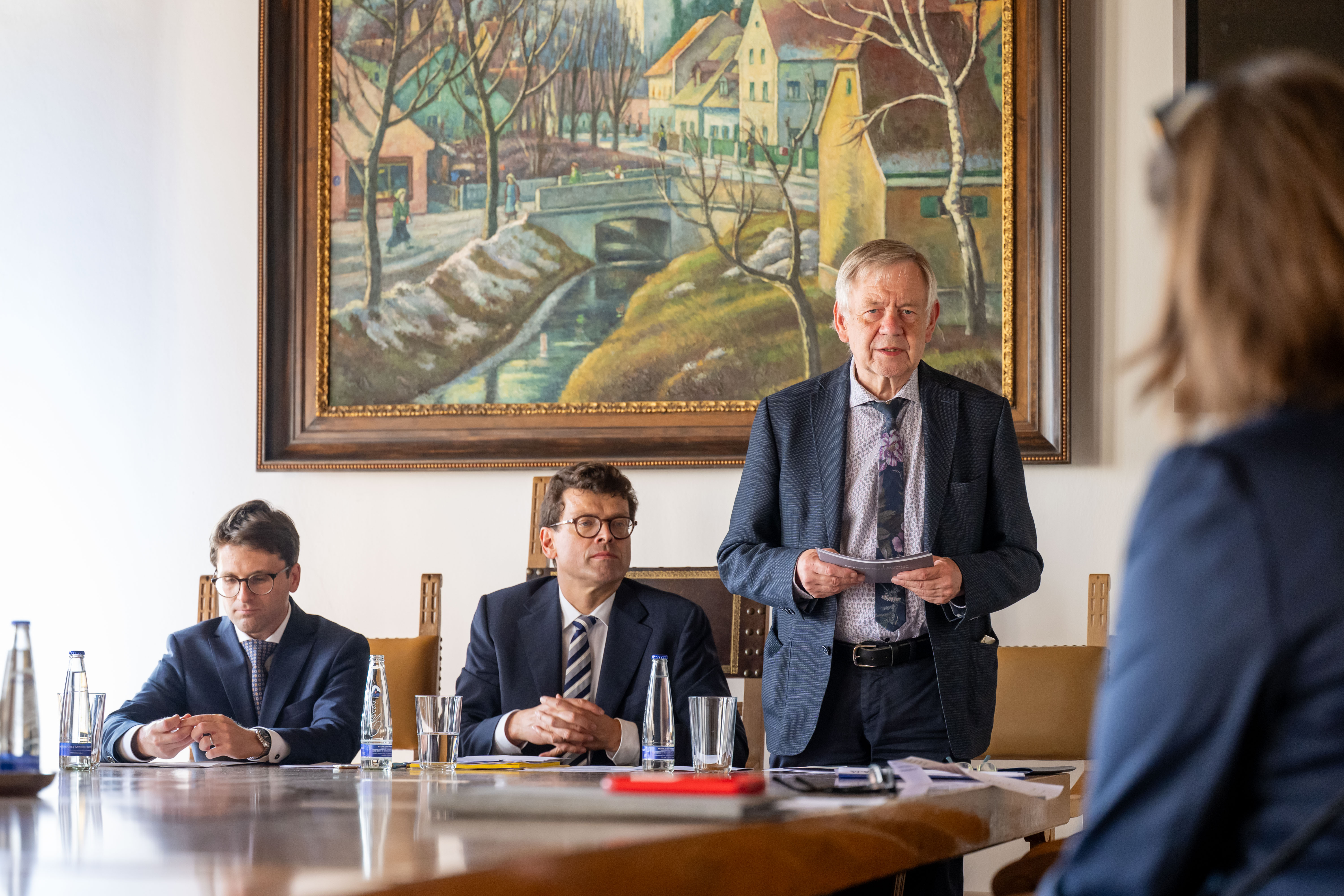Fotografie einer formellen Besprechung: Ein älterer Mann steht mit Unterlagen am Tisch, während zwei Männer sitzen und eine Frau im Vordergrund zuhört. Im Hintergrund hängt ein gerahmtes Landschaftsgemälde; Flaschen, Gläser, Schreibutensilien und Dokumente auf dem Tisch unterstreichen den geschäftlichen Charakter.