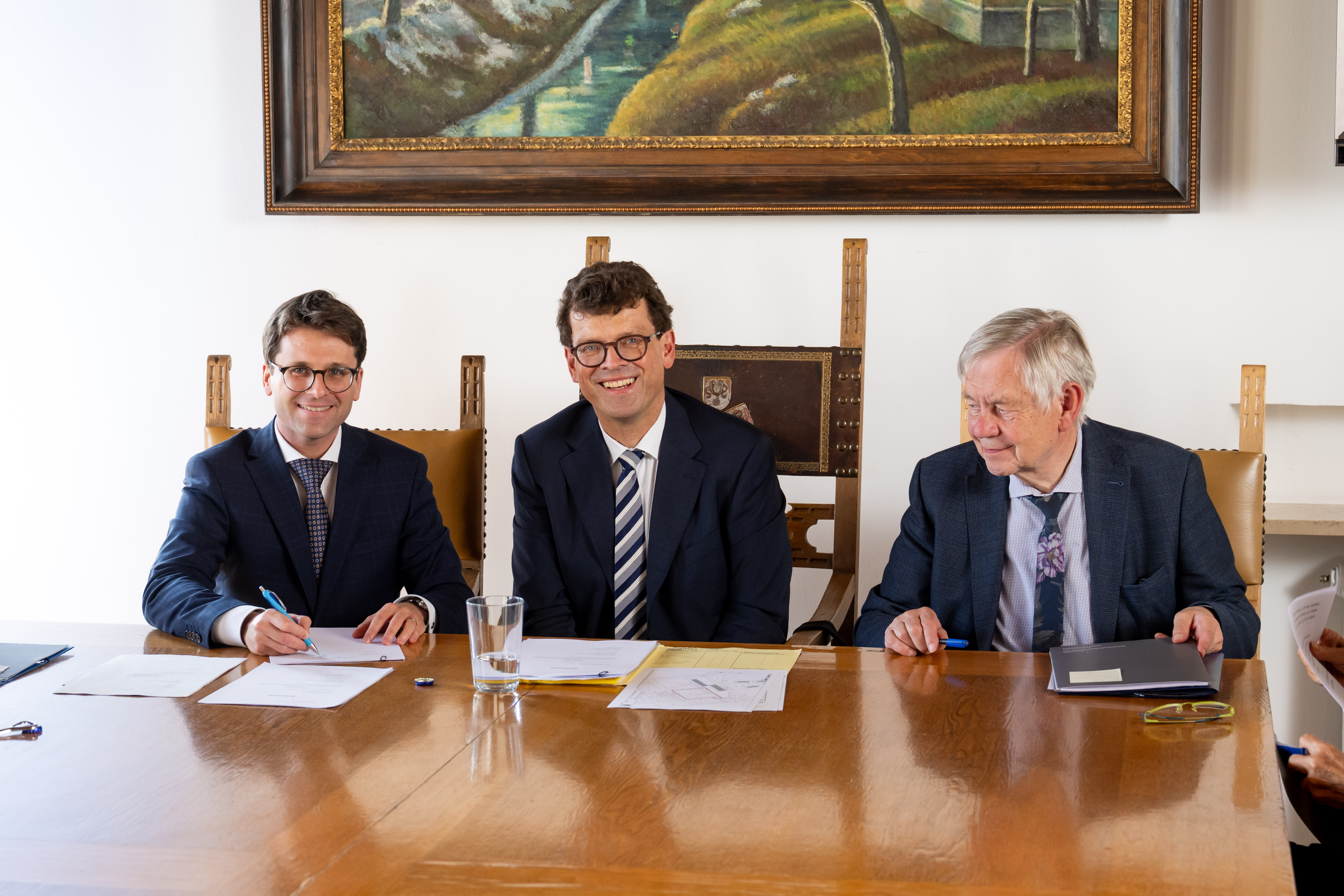 Fotografie von drei formell gekleideten Männern bei einem gemeinsamen Termin an einem Holztisch. Vor ihnen liegen Dokumente und Stifte; links unterschreibt ein Mann, während im Hintergrund ein gerahmtes Landschaftsgemälde hängt.