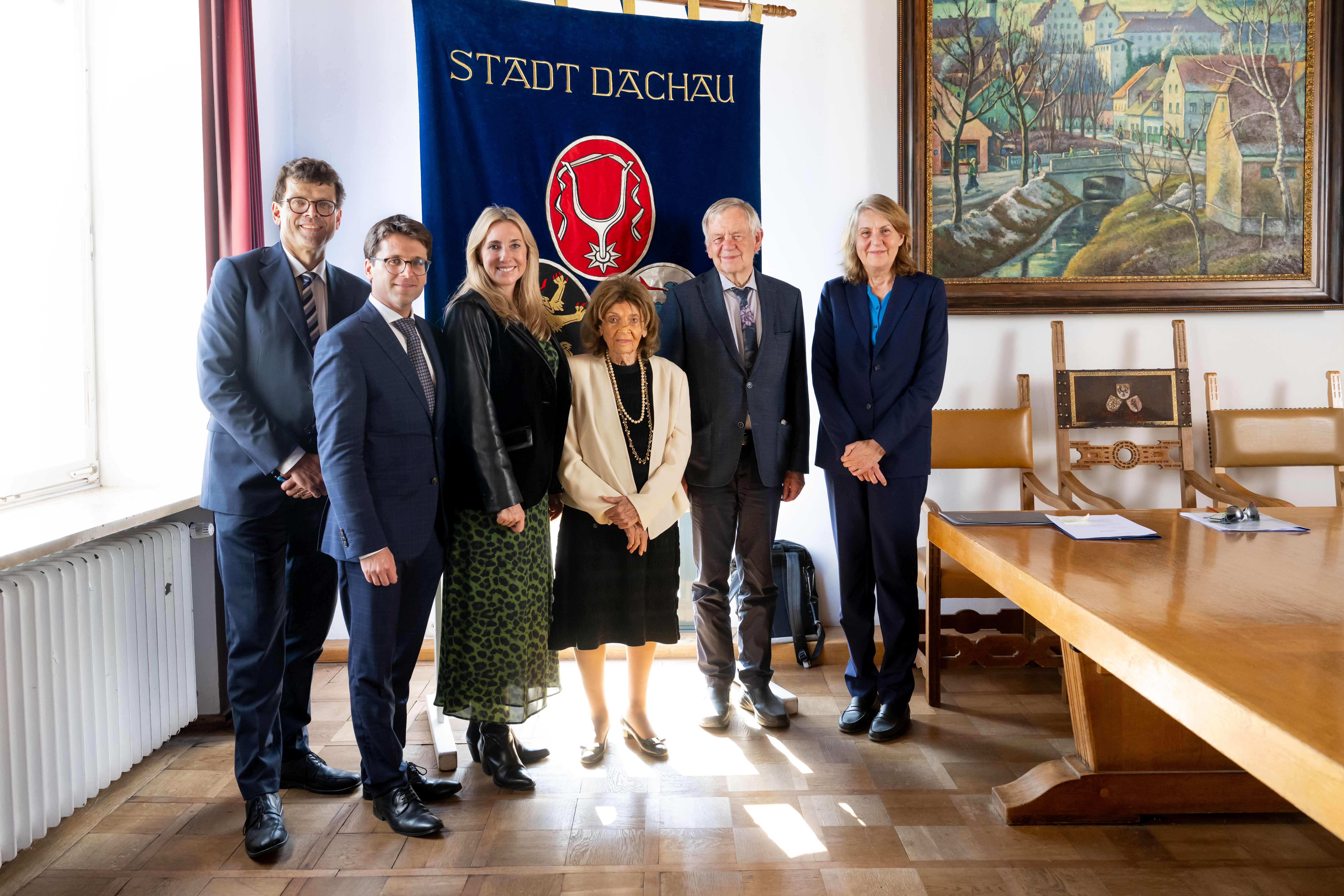 Fotografie von sechs formell gekleideten Personen bei einem offiziellen Termin im Rathaus Dachau. Im Hintergrund hängen ein blaues Banner mit Aufschrift „STADT DACHAU“ und Stadtwappen, ein Gemälde sowie ein Sitzungstisch mit Unterlagen.