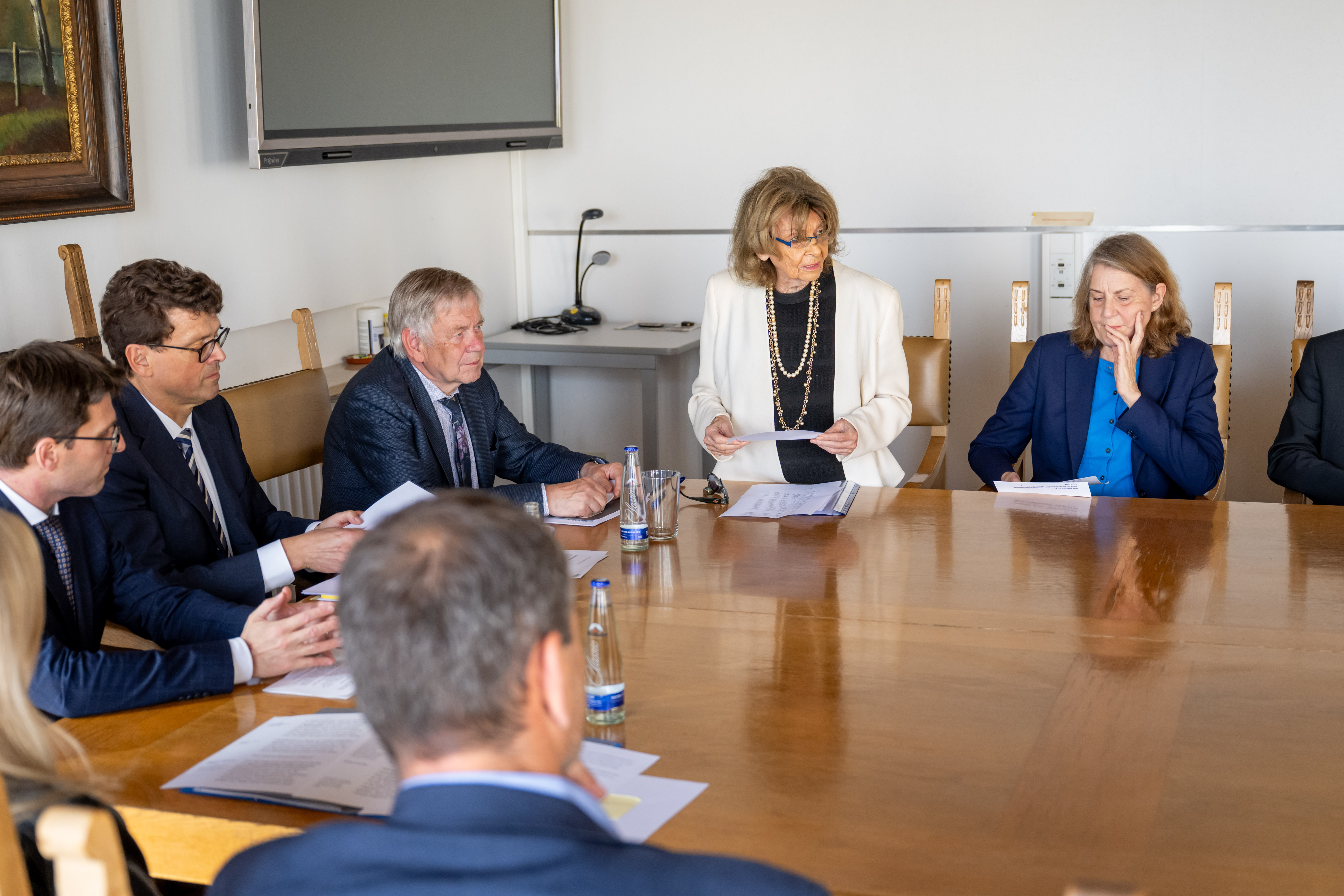 Fotografie einer formellen Besprechung mit mehreren Personen an großem Holztisch. Eine ältere Frau redet gerade zu den Anwesenden Teilnehmern.