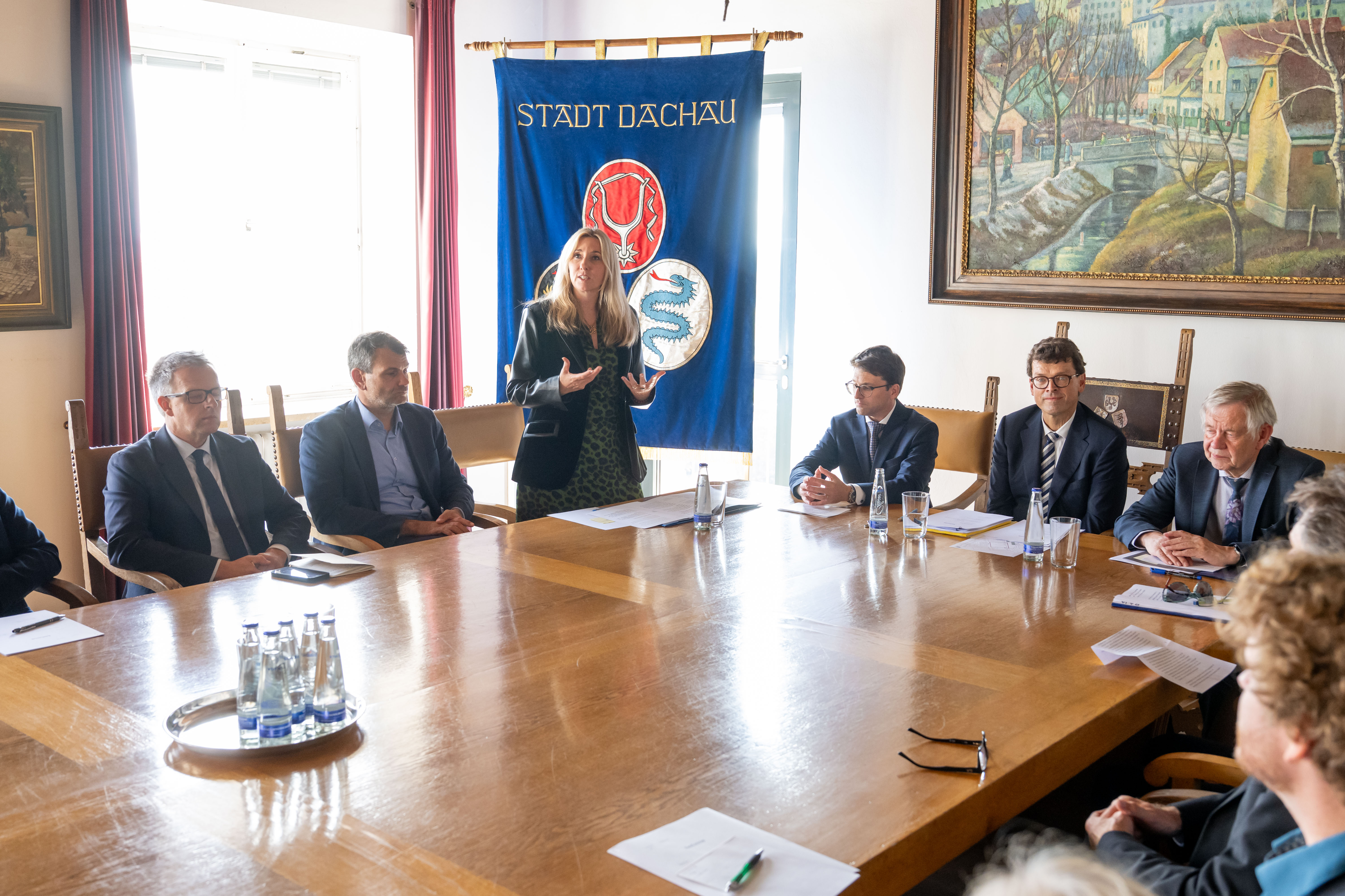 Fotografie einer formellen Besprechung mit mehreren Personen an einem langen Holztisch in repräsentativem Saal. Eine Frau redet gestikulierend