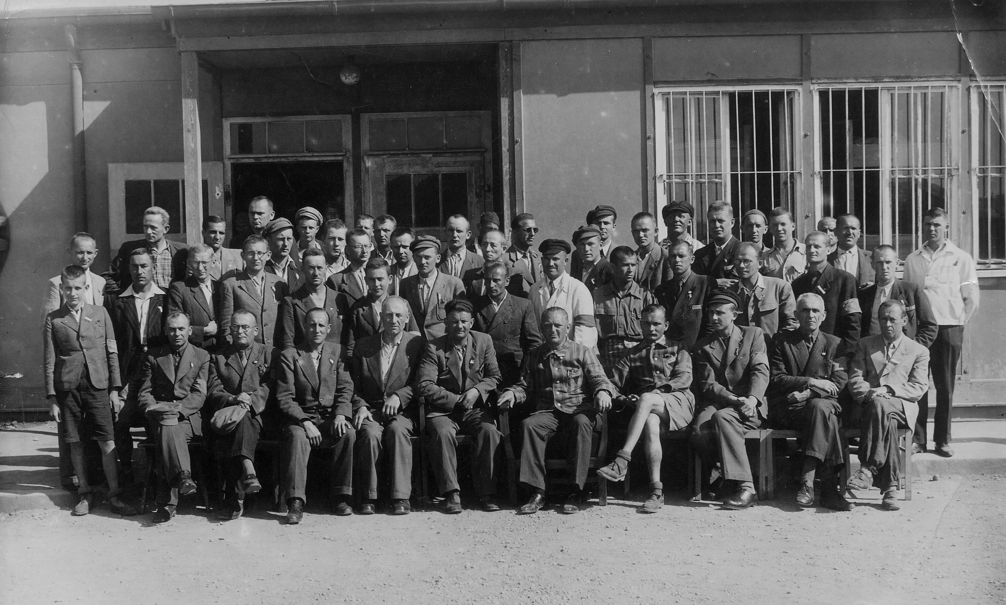 Foto: KZ-Gedenkstätte Dachau Schwarzweiß-Fotografie einer großen Gruppe von Männern, die in mehreren Reihen vor einem Gebäude posieren. Die Männer tragen überwiegend Anzüge oder Arbeitskleidung, einige sitzen auf Stühlen, andere stehen dahinter, was auf ein formelles Gruppenfoto hinweist. Das Foto zeigt eine Gruppe polnischer Häftlinge nach der Befreiung des KZ Dachau.