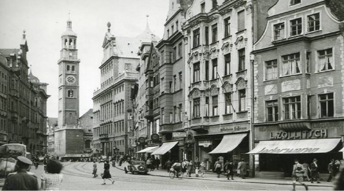 Bild: Filmstill aus dem Film „Die Stille schreit“; zeigt das Haus in der Augsburger Maximilianstraße das von einer Frau Hoffmann „arisiert“ wurde. Frau Hoffmann war 25 Jahre Mitarbeiterin der Augsburger Schirmmanufaktur der Familie Oberdorfer. Die Oberdorfers wurden 1943 deportiert und in Auschwitz ermordet. Die „arische“  Familie Hoffmann feierte im Jahre 1962 ihr „100-jähriges Betriebsjubiläum“