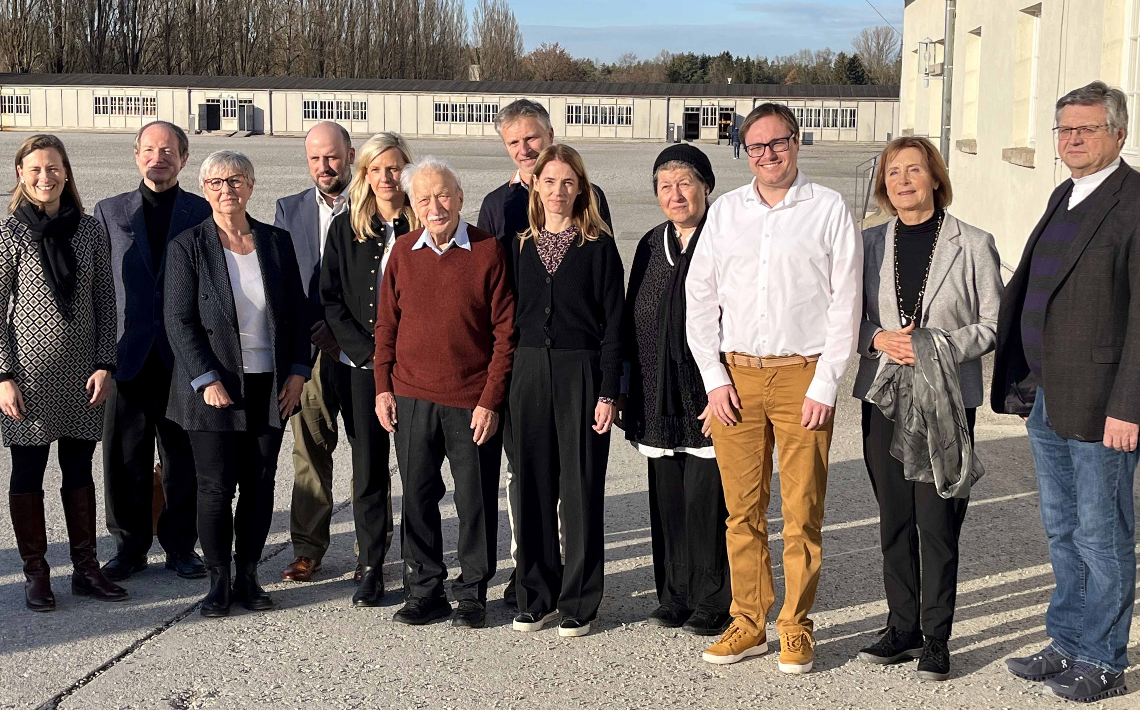 Foto: Stiftung Bayerische Gedenkstätten / G. Hassel Fotografie einer Gruppe von zwölf Personen, die draußen auf einem gepflasterten Platz vor einem langen Gebäude stehen. Die Gruppe zeigt eine Mischung aus Männern und Frauen unterschiedlichen Alters, alle lächelnd und in formeller bis legerer Kleidung