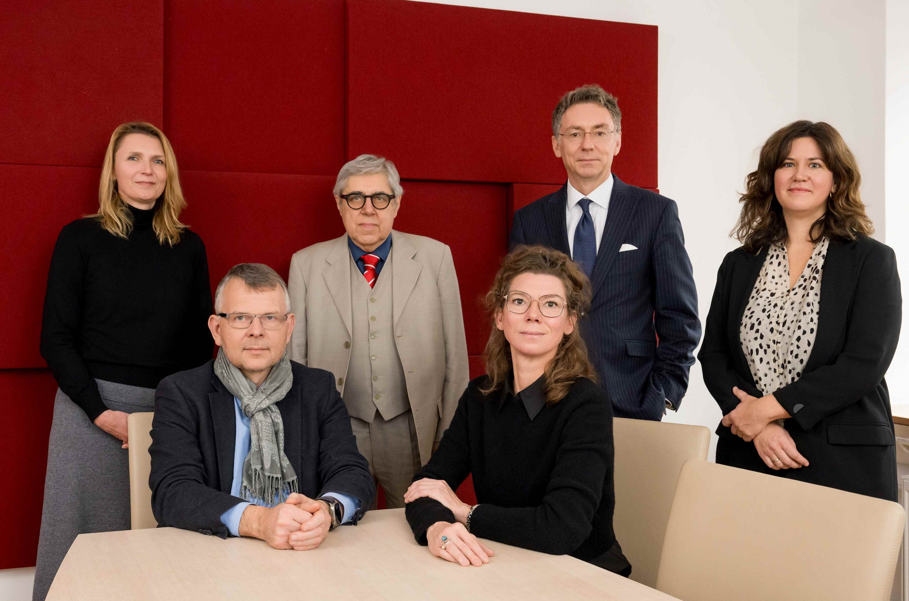 Foto: Stiftung Bayerische Gedenkstätten / T. Hase Gruppenfoto von sechs Personen (drei Männern und drei Frauen)in einem Besprechungsraum mit roter Wandverkleidung, drei Personen sitzen an einem Tisch, drei stehen dahinter. Die Personen tragen formelle Kleidung und blicken in die Kamera.