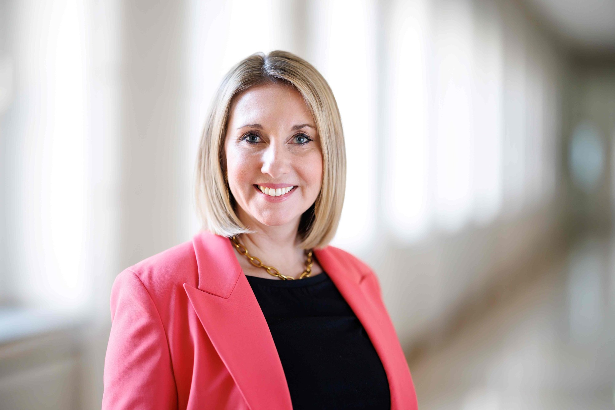 Copyright: StMUK Porträtfoto einer lächelnden Frau mit blonden Haaren, die einen pinken Blazer und eine goldene Halskette trägt. Hintergrund ist unscharf, Fokus auf Gesicht und Oberkörper.