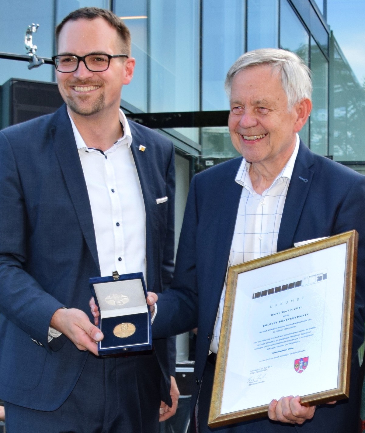 Zwei Männer in Anzügen stehen vor einem modernen Glasgebäude, einer hält eine Medaille in einer Schachtel, der andere eine gerahmte Urkunde. Szene vermittelt Auszeichnung oder Ehrung, beide wirken erfreut und stolz.