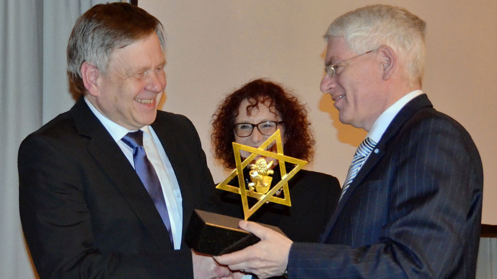 Fotografie zeigt drei Personen bei feierlicher Übergabe einer goldfarbenen Auszeichnung in Form eines Davidsterns auf schwarzem Sockel. Zwei Männer im Anzug übergeben Trophäe, Frau mit Brille und lockigem Haar beobachtet lächelnd im Hintergrund.