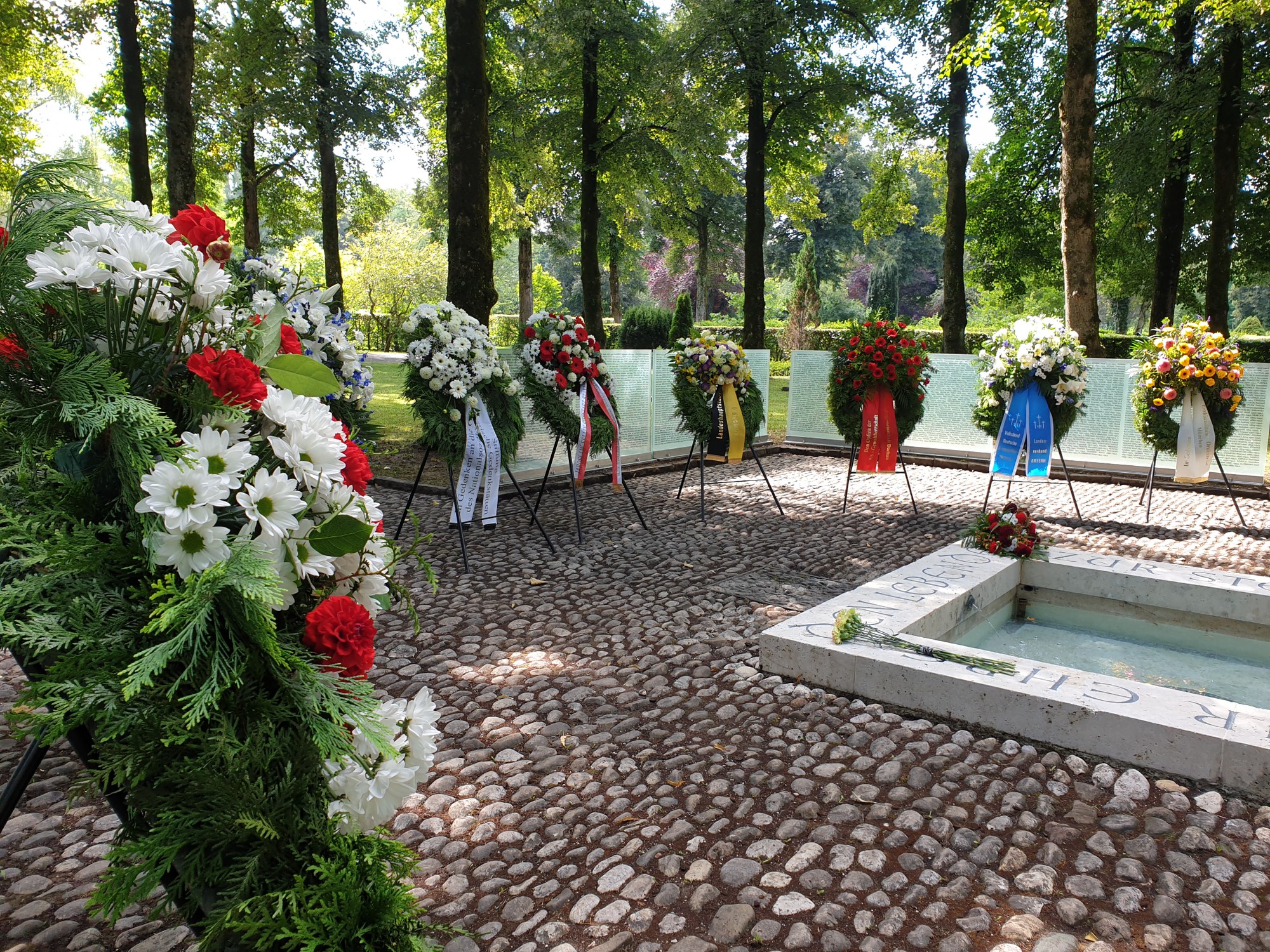 Fotografie eines Gedenkortes mit mehreren Blumenkränzen in verschiedenen Farben, die an einem gepflasterten Bereich und einem Wasserbecken arrangiert sind. Umgeben von Bäumen vermittelt die Szene eine ruhige und respektvolle Atmosphäre, die eine Gedenk- oder Trauerstätte für 3000 Opfer des Nationalsozialismus.