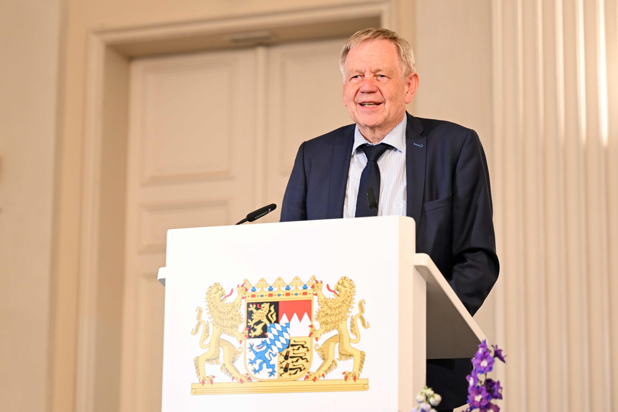 Ein Foto zeigt einen älteren Mann in Anzug und Krawatte, der hinter einem Rednerpult mit bayerischem Wappen steht und spricht. Kontext scheint eine offizielle Rede oder Ansprache in einem formellen Rahmen zu sein.