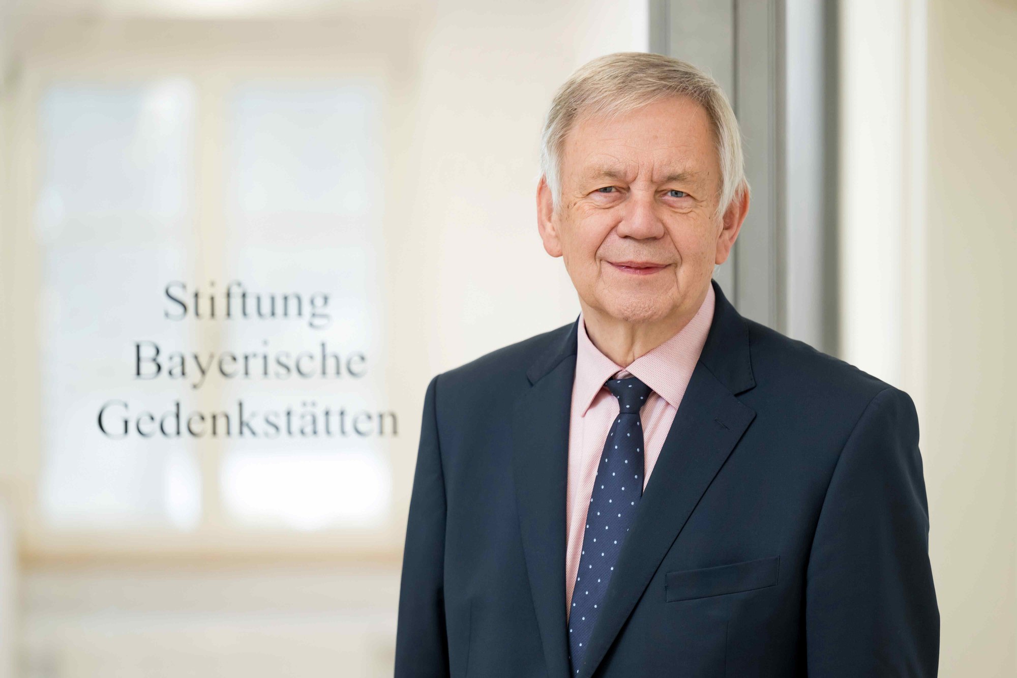 Porträtfoto eines älteren Mannes mit grauen Haaren, der einen dunkelblauen Anzug, rosa Hemd und dunkelblaue Krawatte mit weißen Punkten trägt. Hintergrund unscharf