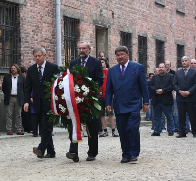 Besuch der Gedenkstätte Auschwitz 2009 Drei Männer in Anzügen tragen einen großen Kranz mit roten und weißen Blumen sowie Bändern durch eine gepflasterte Straße vor Backsteingebäuden. Die Szene wirkt feierlich.