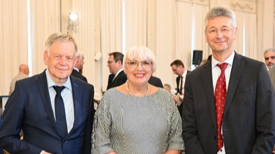Drei Personen in formeller Kleidung stehen in einem hellen Raum mit Vorhängen und mehreren weiteren Personen im Hintergrund. Aufnahme zeigt eine freundliche Atmosphäre bei einer Veranstaltung.