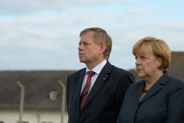 Bundeskanzlerin Dr. A. Merkel bei ihrem ersten Besuch in der KZ-Gedenkstätte Dachau Foto: KZ-Gedenkstätte Dachau Zwei Personen, ein Mann und eine Frau, stehen nebeneinander im Freien vor unscharfem Hintergrund mit Gebäuden. Beide blicken ernst und tragen formelle Kleidung, der Mann mit Anzug und Krawatte, die Frau mit dunklem Blazer.