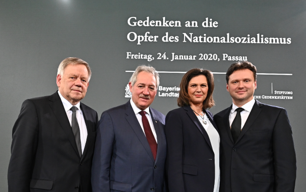 10. gemeinsamer Gedenkakt 2020 in Passau, Foto: Rolf Poss Vier Personen in formeller Kleidung stehen vor einer Wand mit Text zur Gedenkveranstaltung für Opfer des Nationalsozialismus am 24. Januar 2020 in Passau. Die Szene vermittelt einen offiziellen und respektvollen Anlass mit Fokus auf Erinnerung und Mahnung.
