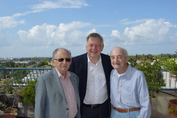 Abba Naor (li.) und Sally Ganor (re.) Drei ältere Männer stehen lächelnd auf einer Dachterrasse mit Blick auf eine Stadtlandschaft und blauen Himmel. Die Holocaust-Überlebenden Abba Naor (links) und Solly Ganor (rechts) haben sen Stiftungsdirektor in ihrer Mitte