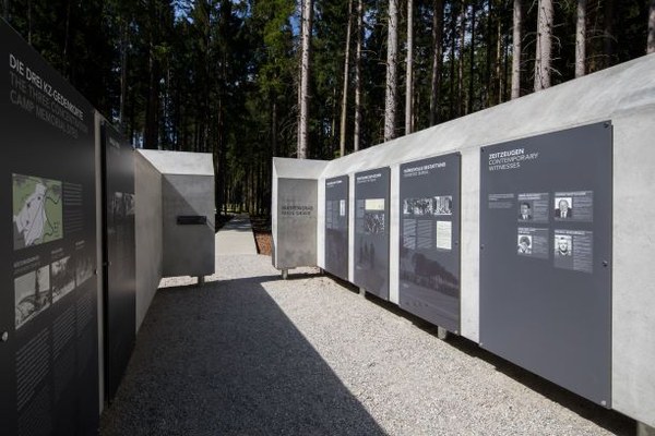 Mühldorfer Hart (2018) Foto: Heiner Heine, Burgkirchen Außenbereich mit mehreren großen, dunkelgrauen Informationstafeln, die entlang eines Kieswegs zwischen Betonwänden aufgestellt sind. Tafeln enthalten Text, Fotos und Karten, und sind Teil einer Gedenkstätte.
