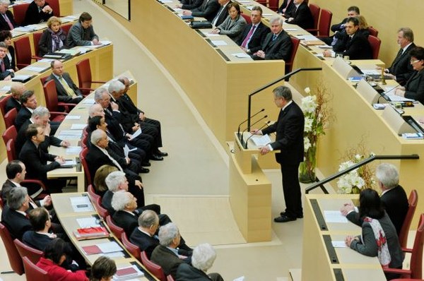Erster gemeinsamer Gedenkakt im Bayerischen Landtag 2011 Fotografie mit einem Redner am Rednerpult und mehreren sitzenden Abgeordneten in einem halbkreisförmigen Saal im Plenum des Bayerischen Landtags.