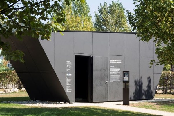 Fotografie eines modernen, schwarzen Kubus mit geöffnetem Eingang, der als Gedenkstätte und Ausstellung dient. Beschriftungen in weiß an der Fassade geben Informationen, umgeben von Bäumen und Grünfläche.
