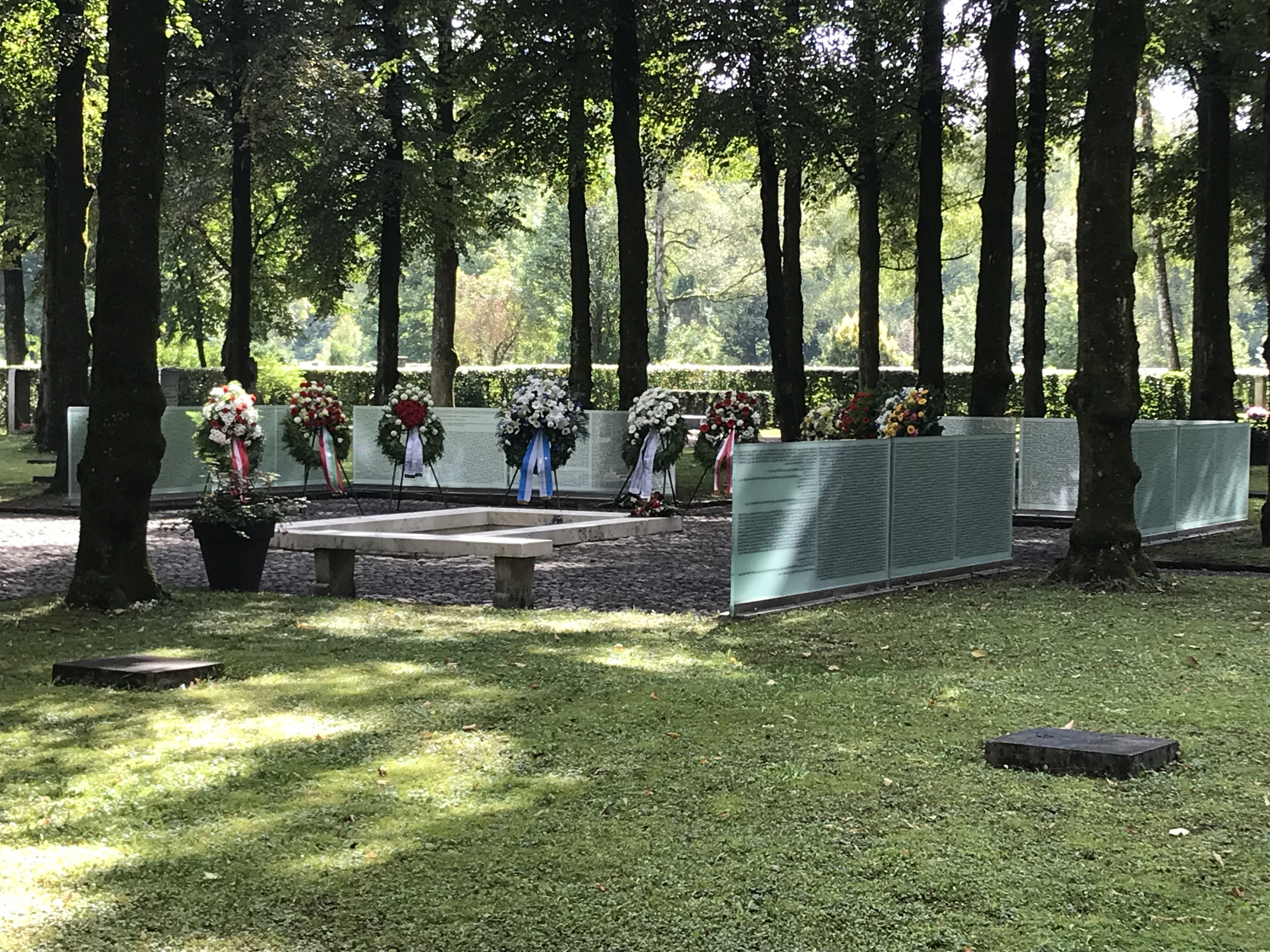 Foto:SBG Fotografie eines Gedenkorts mit Glaswänden, auf denen Namen eingraviert sind, umgeben von Bäumen und einem Rasen. Mehrere Blumengestecke stehen als Zeichen der Erinnerung an einer Wand, während eine Bank zum Verweilen einlädt.