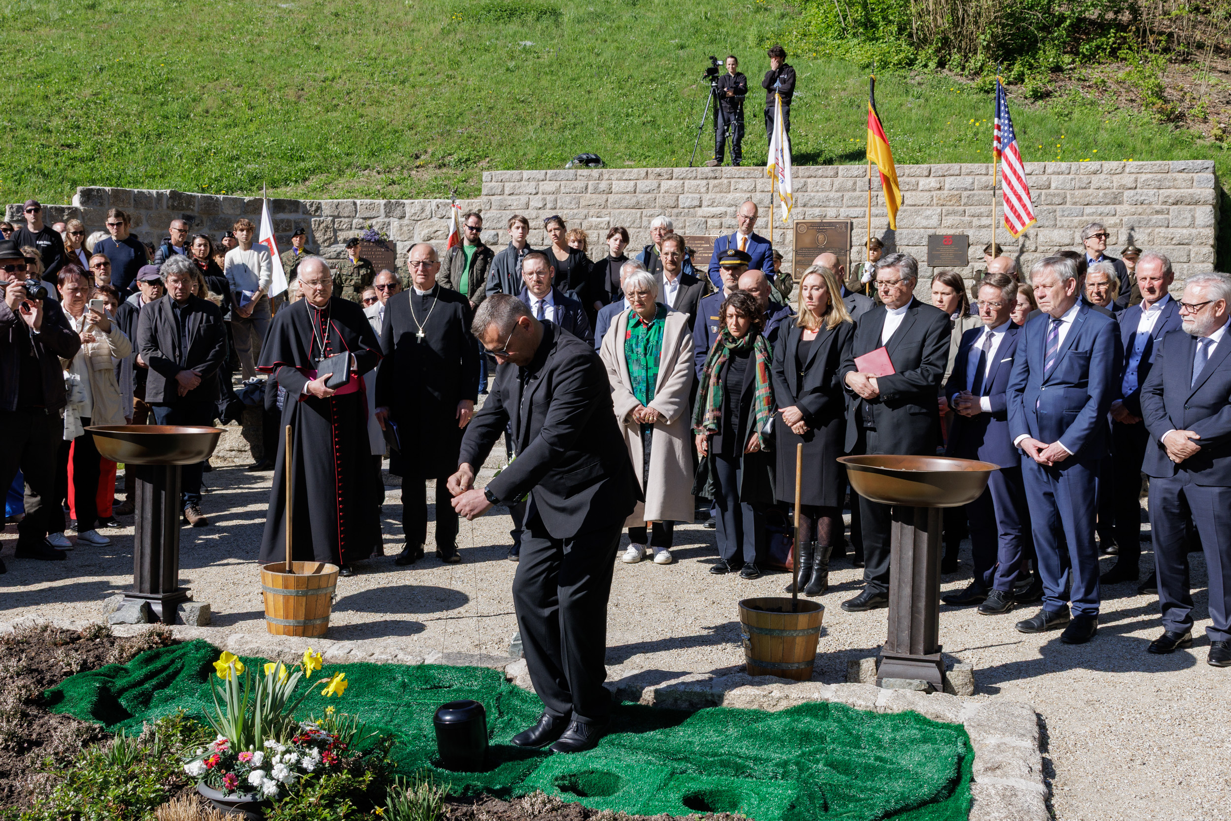 Foto: KZ-Gedenkstätte Flossenbürg / T. Dashuber Fotografie einer Gedenkzeremonie mit einer großen Gruppe von Menschen, darunter Geistliche und Offizielle, die vor einem Urnengrab versammelt sind. Im Vordergrund legt ein Geistlicher die Urne in das vorbereitete Grab.