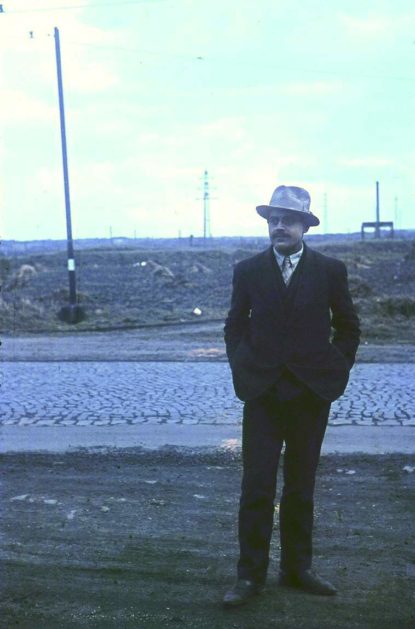 Copyright: Federal Archives Berlin Photograph of a man in a suit and hat standing on unpaved ground in front of a cobbled street. In the background, a mast and a vast, barren landscape with scattered structures can be seen.