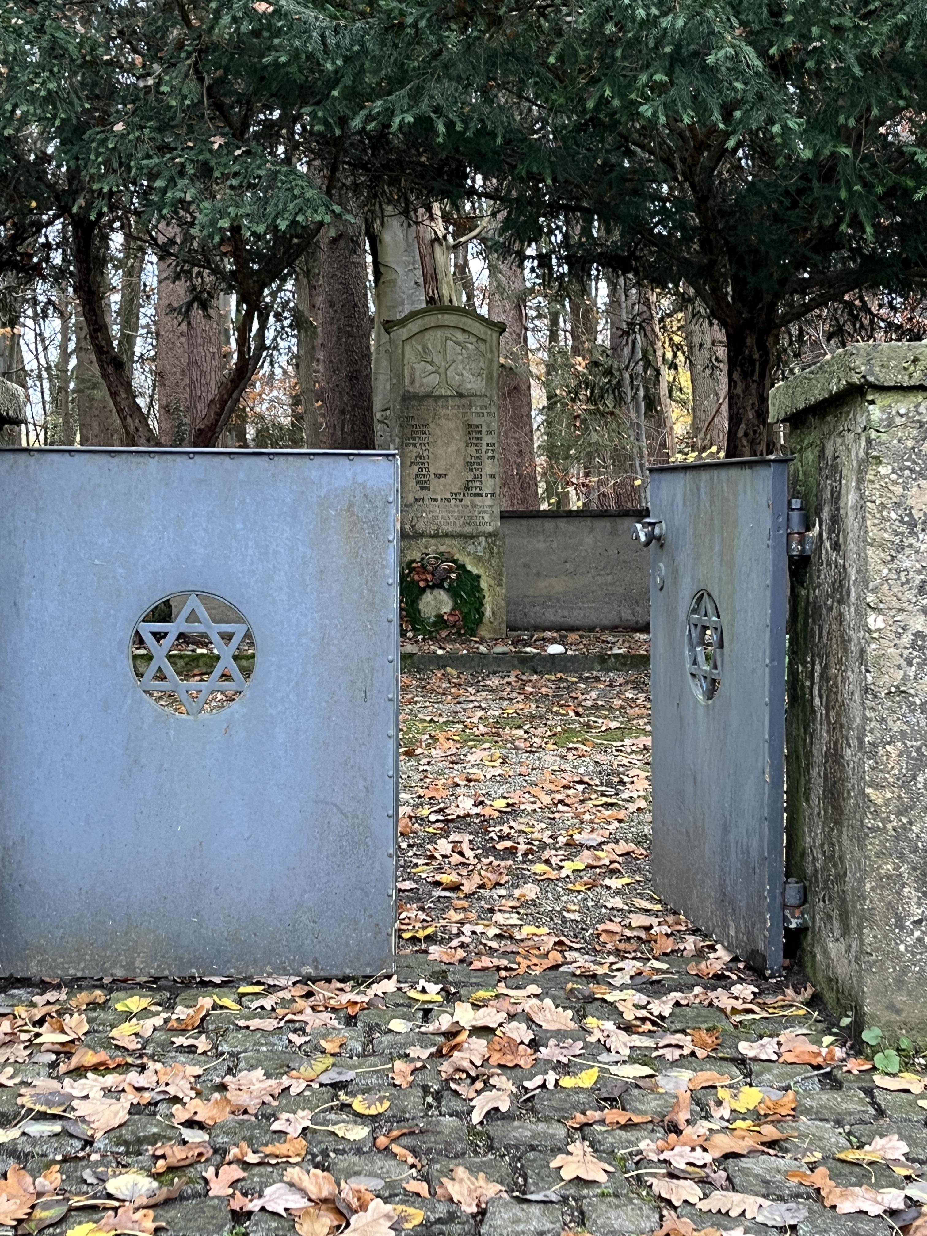 Offenes blaues Tor mit Davidsternen führt zu einem Gedenkstein auf einem mit Herbstlaub bedeckten Friedhof