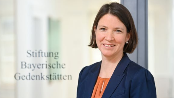 Porträtfotografie einer Frau mit braunen Haaren, die in einem orangen Oberteil und dunkelblauen Blazer lächelnd vor einem hellen Hintergrund steht. Links neben ihr steht der Text „Stiftung Bayerische Gedenkstätten