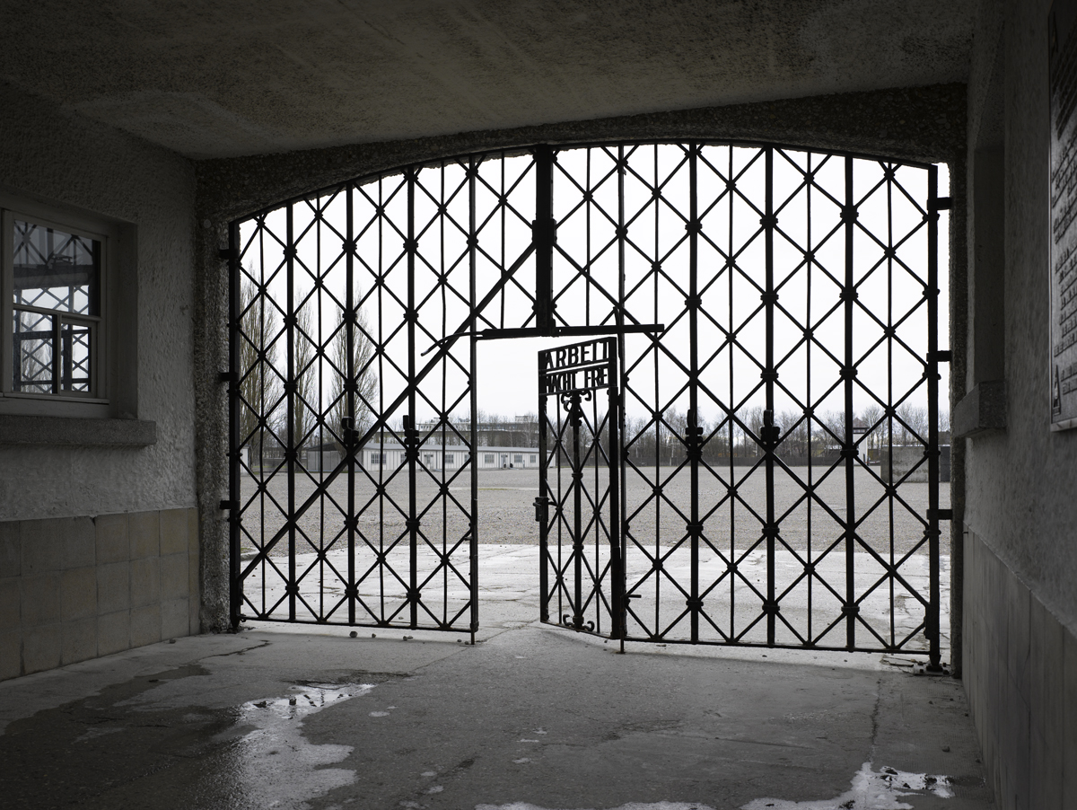 Ehemaliges Lagertor Copyright: KZ-Gedenkstätte Dachau / S.Müller-Naumann Fotografie eines weitläufigen, symmetrisch angeordneten Gebäudekomplexes mit weiß getünchten Mauern und dunklen Dächern, eingerahmt von zwei großen Bäumen im Vordergrund. Bewölkter Himmel und ruhige Atmosphäre betonen die historische Bedeutung des Ortes.