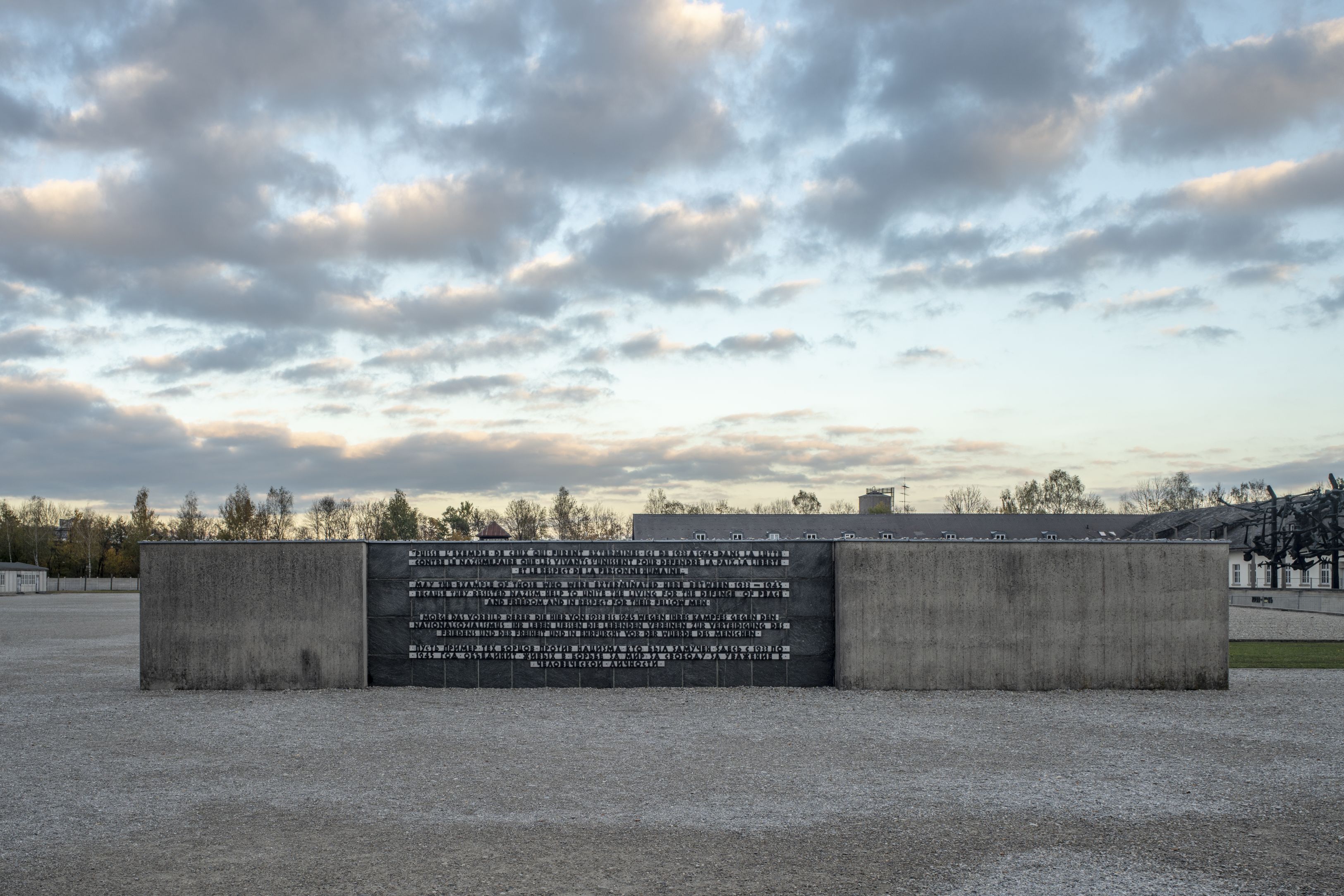 Internationales Mahnmal Copyright KZ-Gedenkstätte Dachau / Rainer Viertlböck Fotografie eines modernen Denkmals aus Beton mit einer schwarzen Tafel, auf der Text eingraviert ist, vor einem bewölkten Himmel. Das Denkmal steht auf einem gepflasterten Platz .