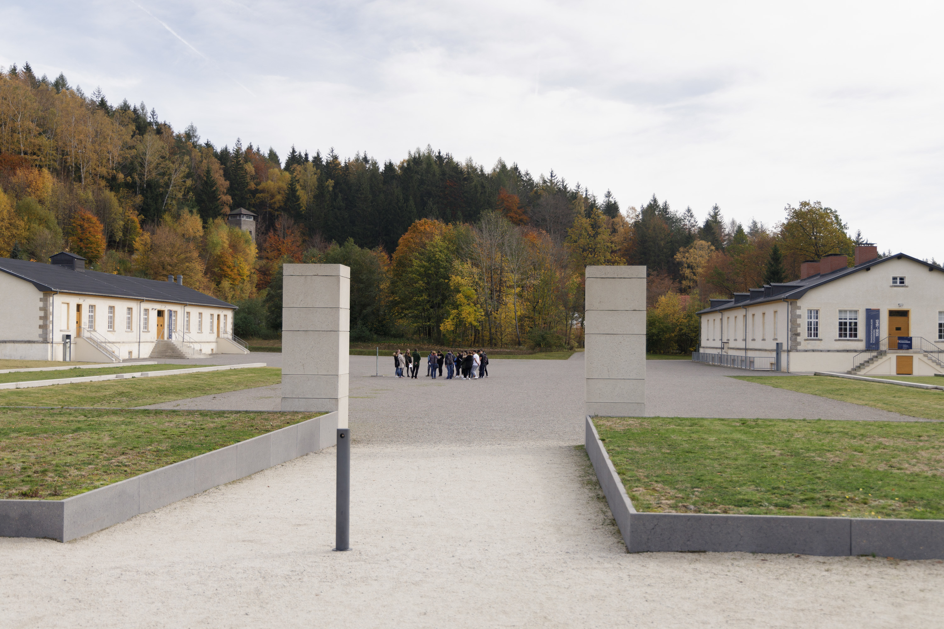 Lagertor und Appellplatz Foto KZ-Gedenkstätte Flossenbürg / Th. Dashuber Fotografie eines historischen Gedenkorts mit zwei niedrigen Gebäuden links und rechts, umgeben von herbstlich gefärbtem Wald. Im Vordergrund ein offener Bereich mit Betonwänden und einem kleinen Gruppen von Menschen in der Bildmitte. Das Bild zeigt den ehemaligen Appellplatz in der KZ-Gedenkstätte Flossenbürg.