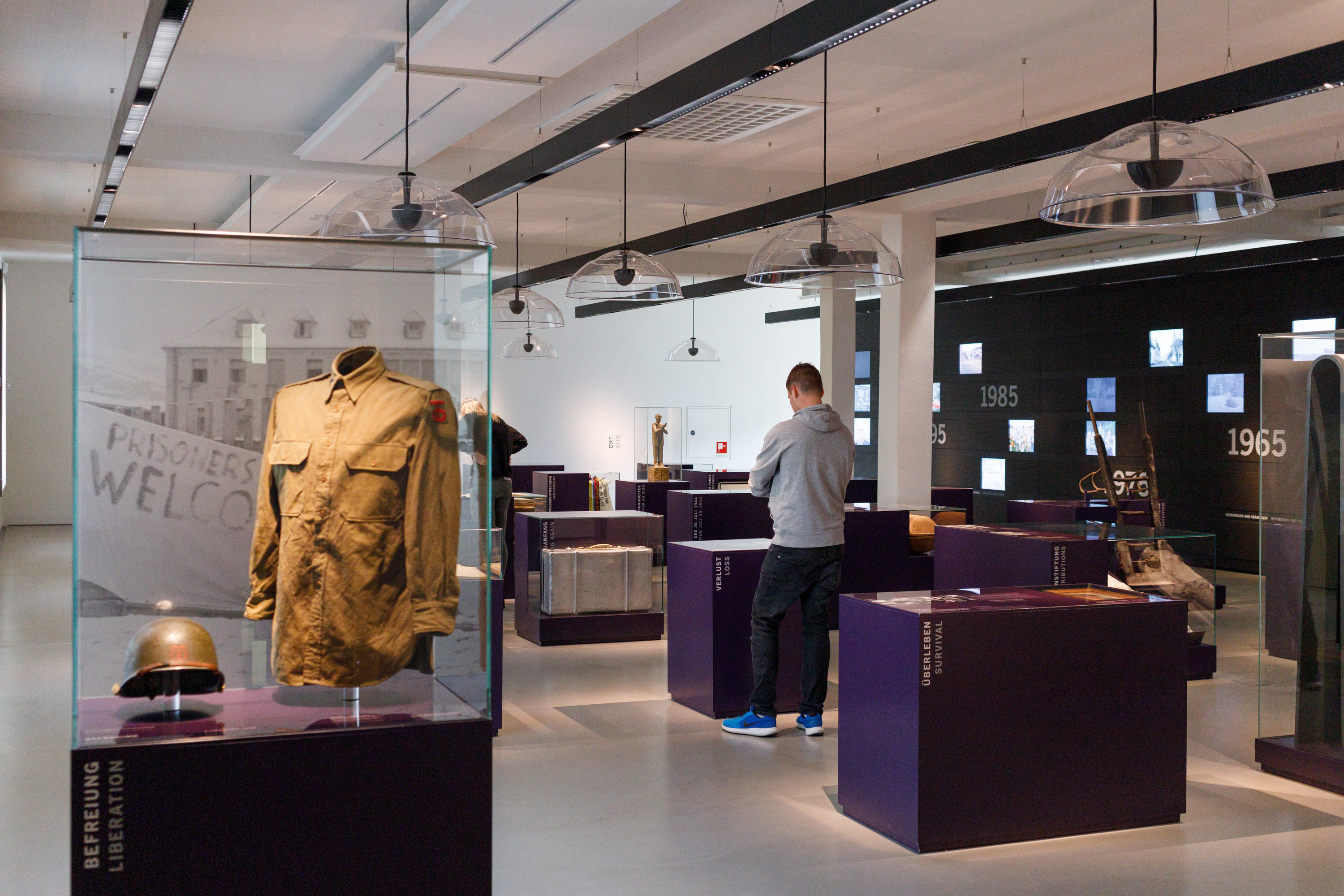 Was bleibt Foto KZ-Gedenkstätte Flossenbürg / Th. Dashuber Fotografie eines Museumsraums mit Ausstellungsstücken, darunter im Vordergrund eine Jacke und ein Helm in einer Glasvitrine. Besucher betrachten Exponate auf violetten Podesten, während eine beleuchtete Wand im Hintergrund Jahreszahlen und Informationen zeigt. Das Bild zeigt einen Blick in die Ausstellung der KZ-Gedenkstätte Flossenbürg.