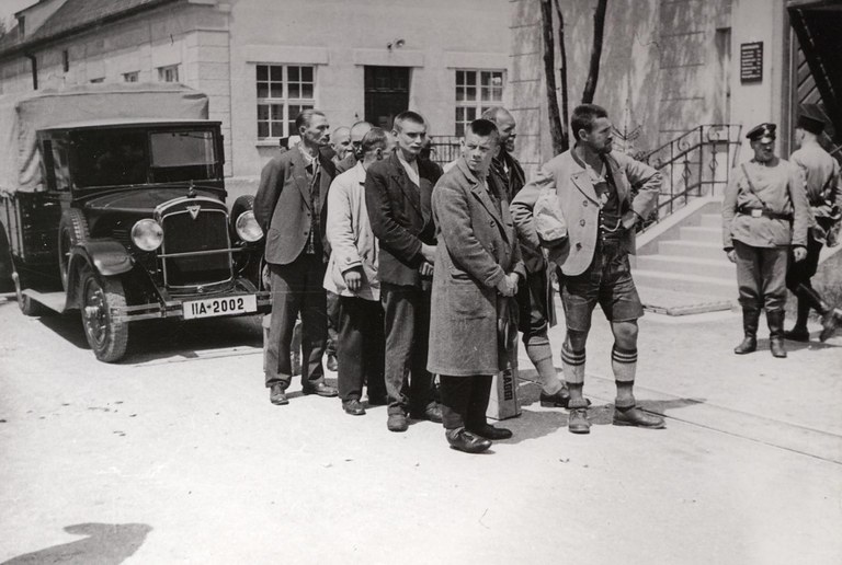Vor einem Auto stehen acht Häftlinge stehen in zweier Reihe hintereinander. Neben ihnen sind 2 uniformierte Offiziere (Polizei) zu sehen