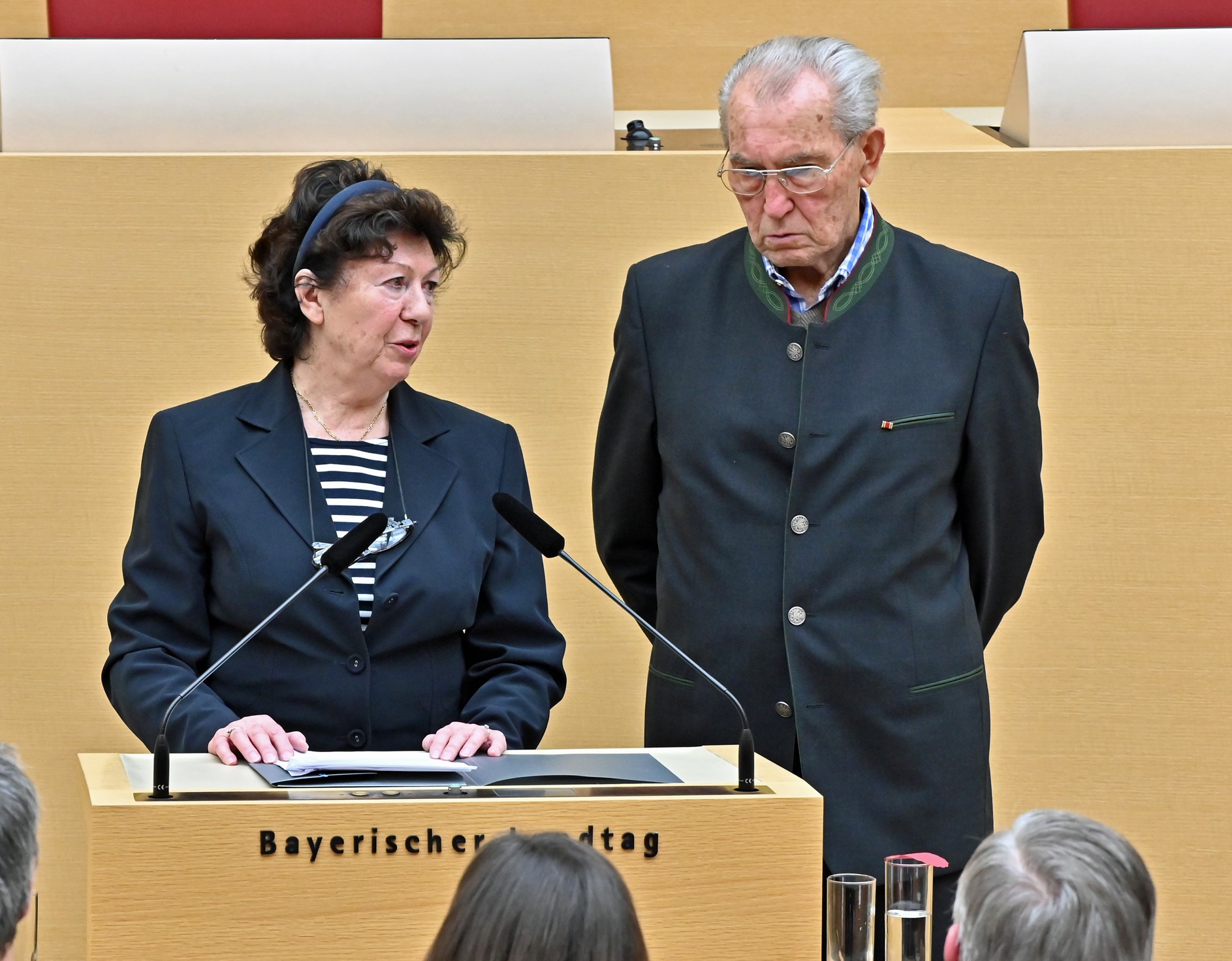 Schwarzweißfotografie aus dem Bayerischen Landtag: Eine Frau im blauen Anzug spricht am Rednerpult, während ein älterer Mann im Janker neben ihr steht. Zwei Mikrofone, Unterlagen, Gläser und sitzende Zuhörende im Vordergrund verdeutlichen eine Redessituation. Am Rednerpult steht "Bayerischer Landtag".
