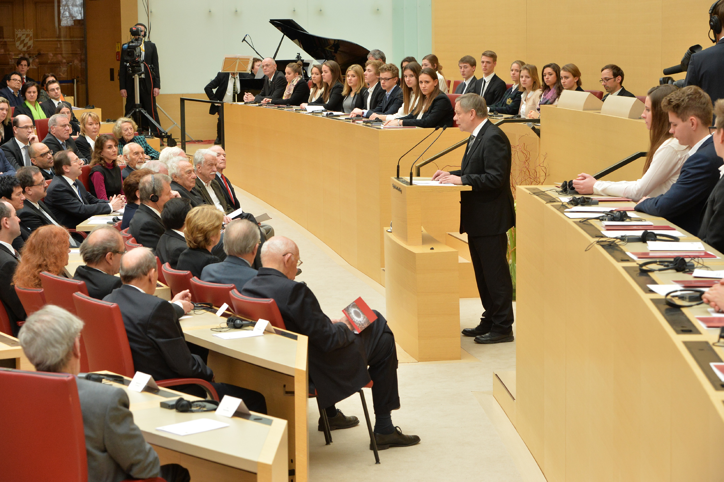 Foto: Bayerischer Landtag / Rolf Poss Fotografie einer formellen Versammlung in einem Sitzungssaal im Bayerischen Landtag mit einem Redner am Podium und aufmerksam zuhörendem Publikum. Es sind mehrere Personen in Anzügen und einem Orchester im Hintergrund zu sehen.