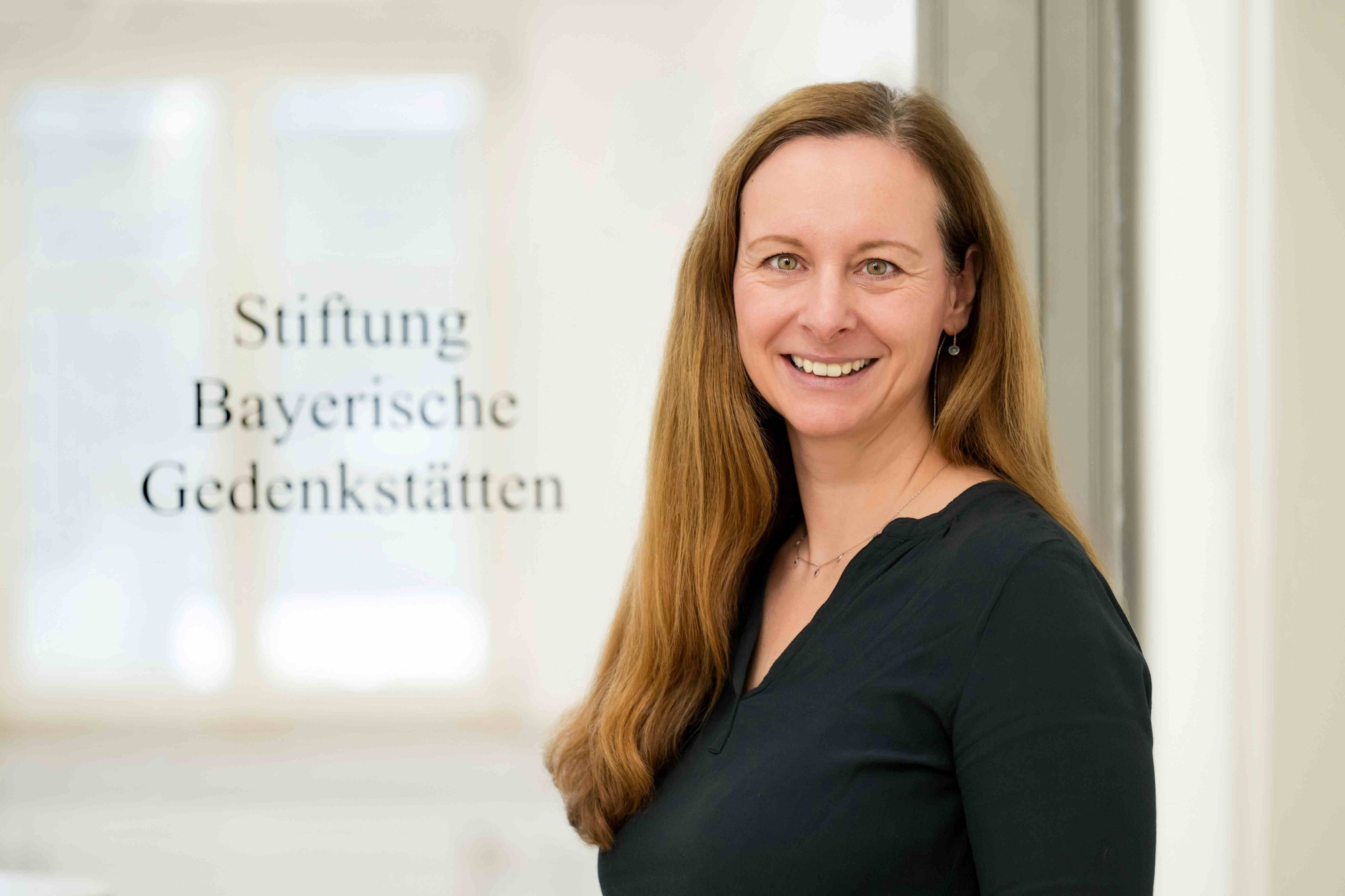 Porträtfoto einer lächelnden Frau mit langen braunen Haaren vor unscharfem Hintergrund mit Schriftzug „Stiftung Bayerische Gedenkstätten“.