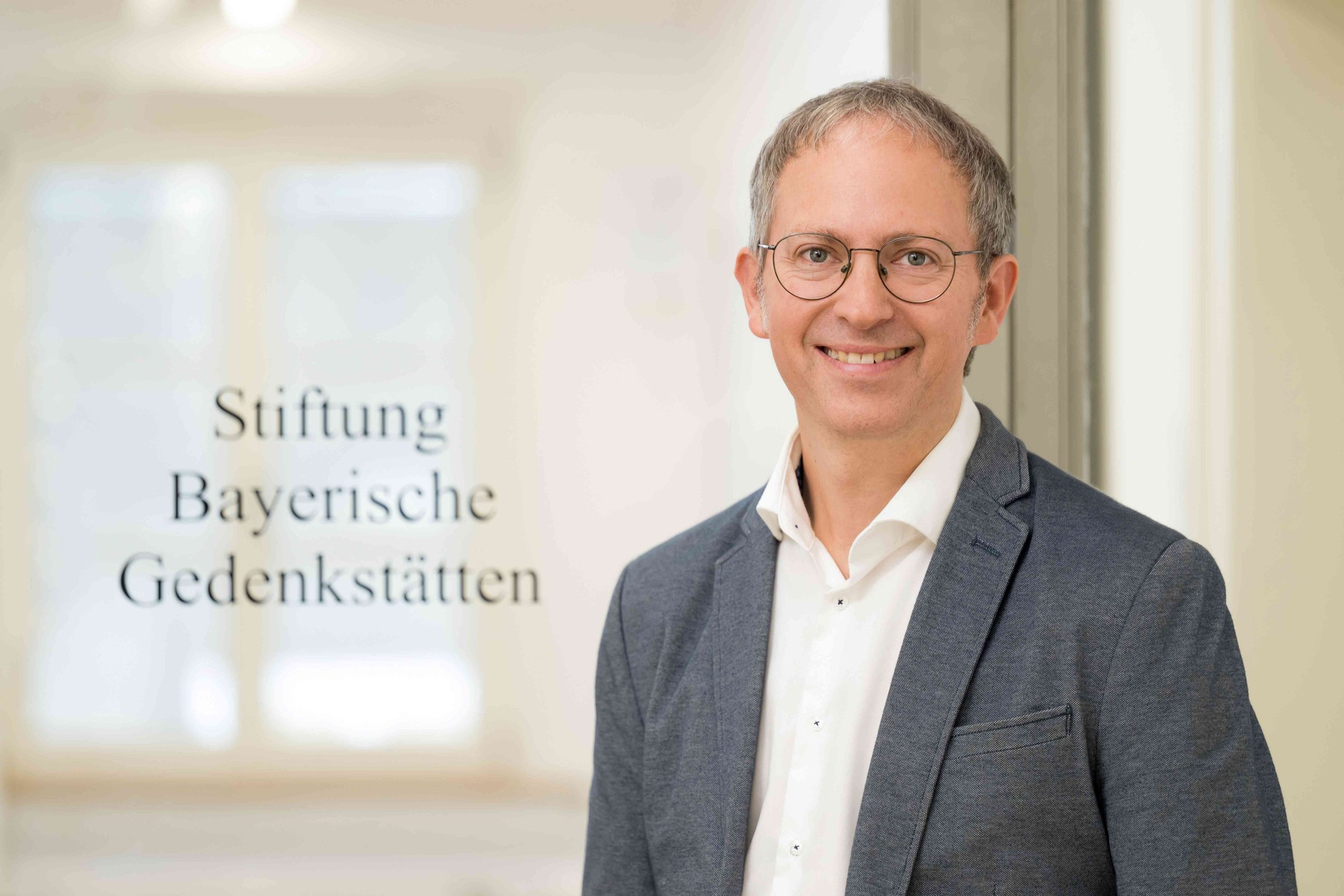 Porträtfotografie eines lächelnden Mannes mit Brille und grauem Sakko vor einem Fenster mit Schriftzug „Stiftung Bayerische Gedenkstätten“.