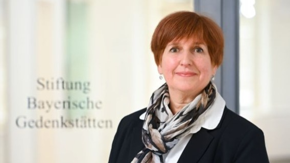 Porträtfotografie einer Frau mit kurzem roten Haar, schwarzem Blazer und gemustertem Schal vor einer Glaswand mit der Aufschrift "Stiftung Bayerische Gedenkstätten". 