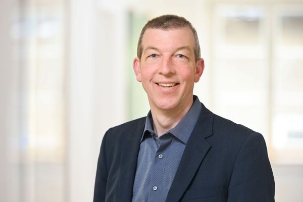 Porträtfoto eines lächelnden Mannes in Business-Kleidung vor unscharfem, hellem Hintergrund. 
