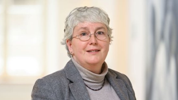 Porträtfotografie einer Frau mit Brille und angegrauten Haaren, in einem grauen Rollkragenpullover und und dunkelgrauem Blazer 