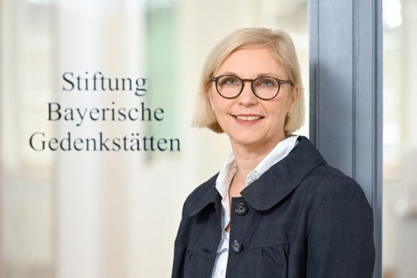 Porträtfoto einer lächelnden Frau mit Brille vor einer Glaswand mit Schriftzug "Stiftung Bayerische Gedenkstätten"
