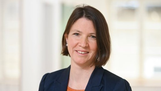 Porträtfotografie einer Frau mit braunen Haaren, die in einem orangen Oberteil und dunkelblauen Blazer lächelnd vor einem hellen Hintergrund steht. 