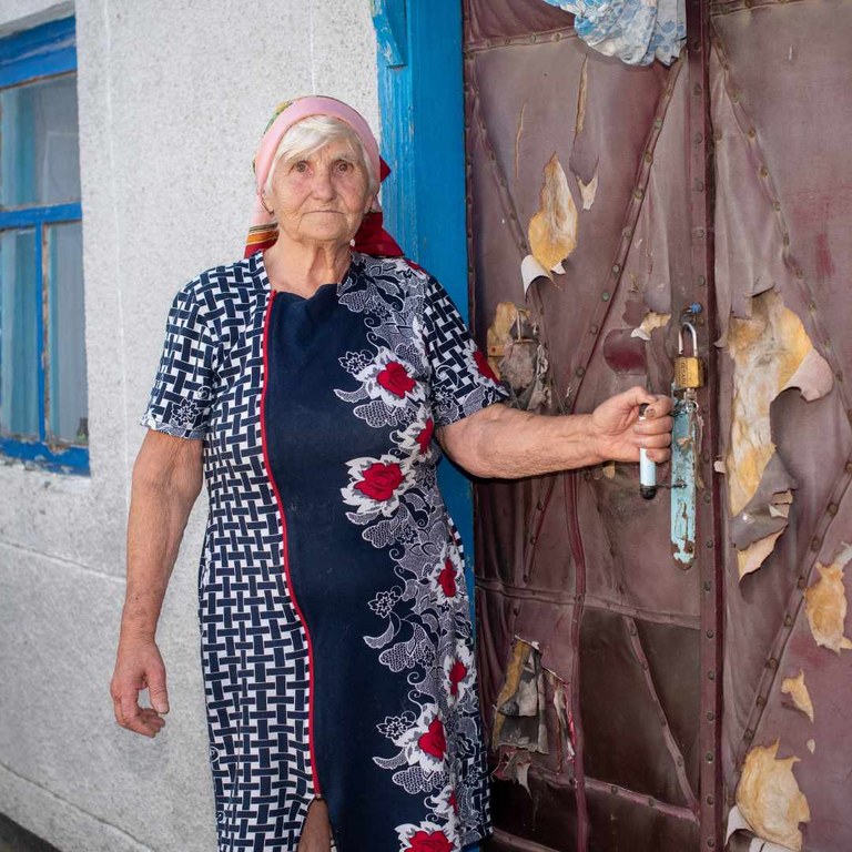 Eine alte Frau mit rotem Kopftuch steht an einer zerschlissenen, mit Sicherheitsschloss versehenen Haustür