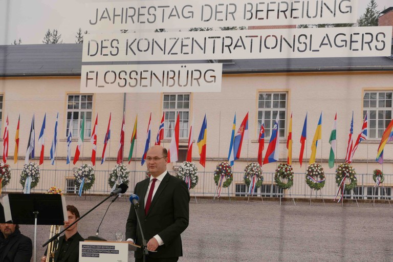 Der Staatsminister steht vor einem Bild mit vielen Fahnen und dem Schriftzug "Jahrestag der Befreiung des Konzentrationslagers Flossenbürg"