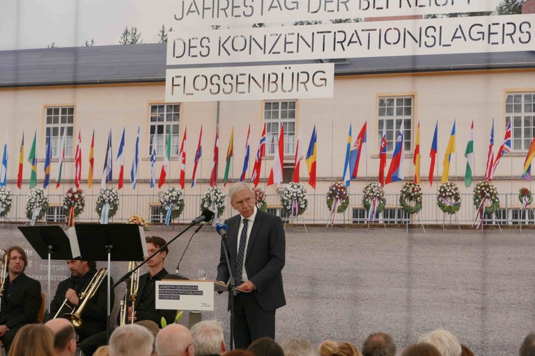 Der Prof. steht vor einem Bild mit vielen Fahnen und dem Schriftzug "Jahrestag der Befreiung des Konzentrationslagers Flossenbürg"