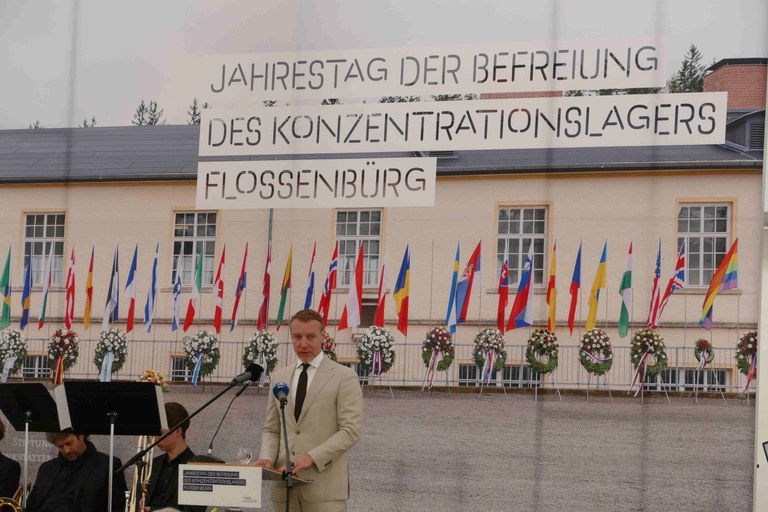 Er steht vor einem Bild mit vielen Fahnen und dem Schriftzug "Jahrestag der Befreiung des Konzentrationslagers Flossenbürg"