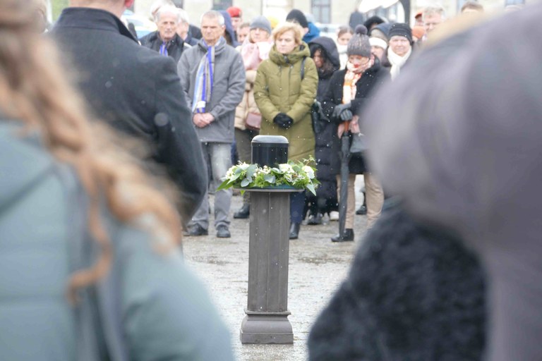 eine schwarze Urne steht von einem Kranz mit weißen Blumen umrandet auf einem Prodest. Die Gäste haben sich darum aufgestellt.