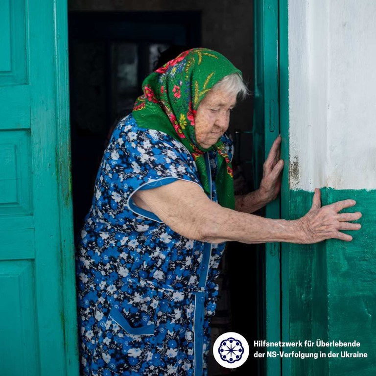 Eine alte Frau mit grünem Kopftuch öffnet die Haustür